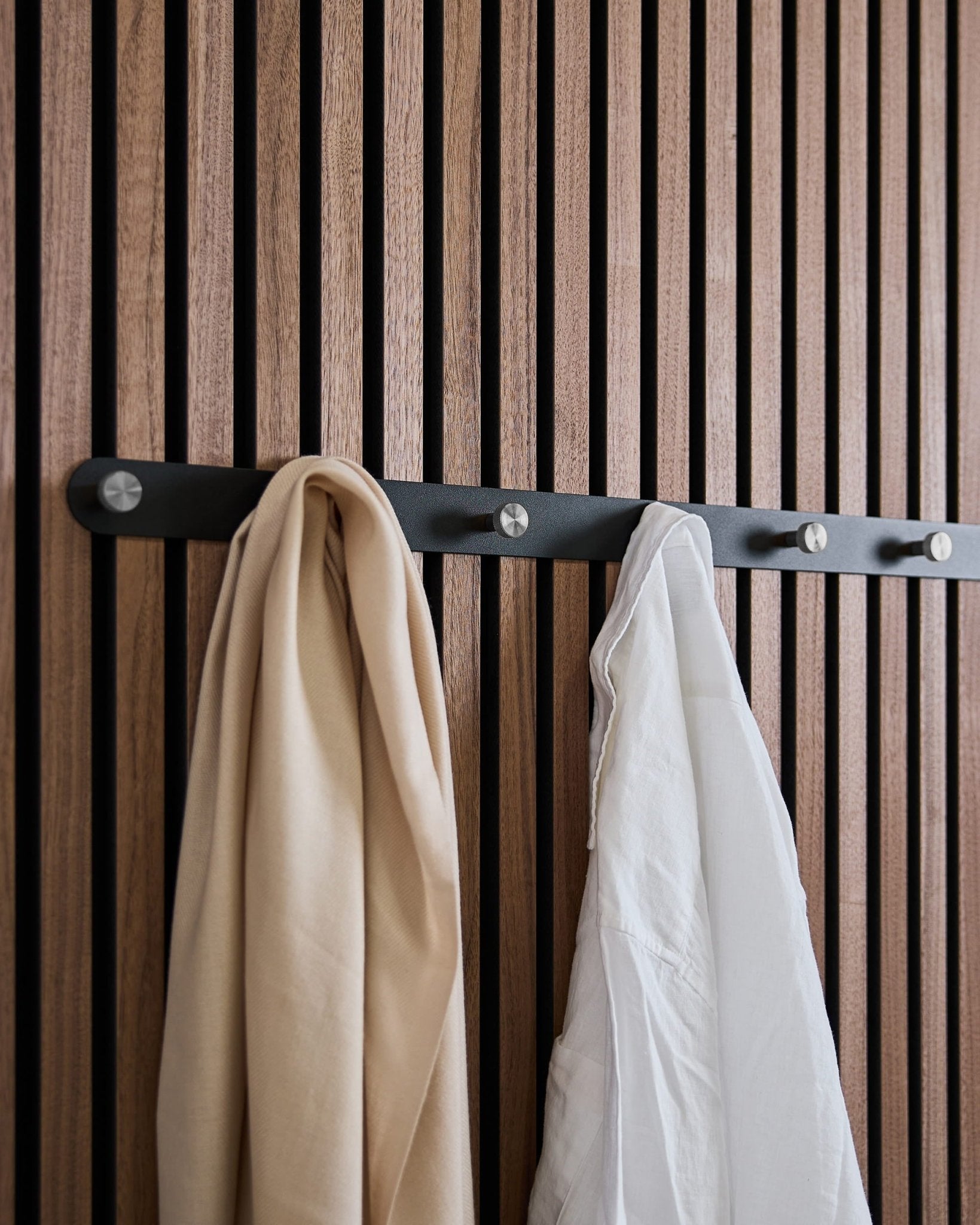 A beige scarf and white shirt hang on WoodUpp’s Norr X7 Black/Stainless Steel coat hook rack, mounted to a Nordic acoustic slatwall panel.